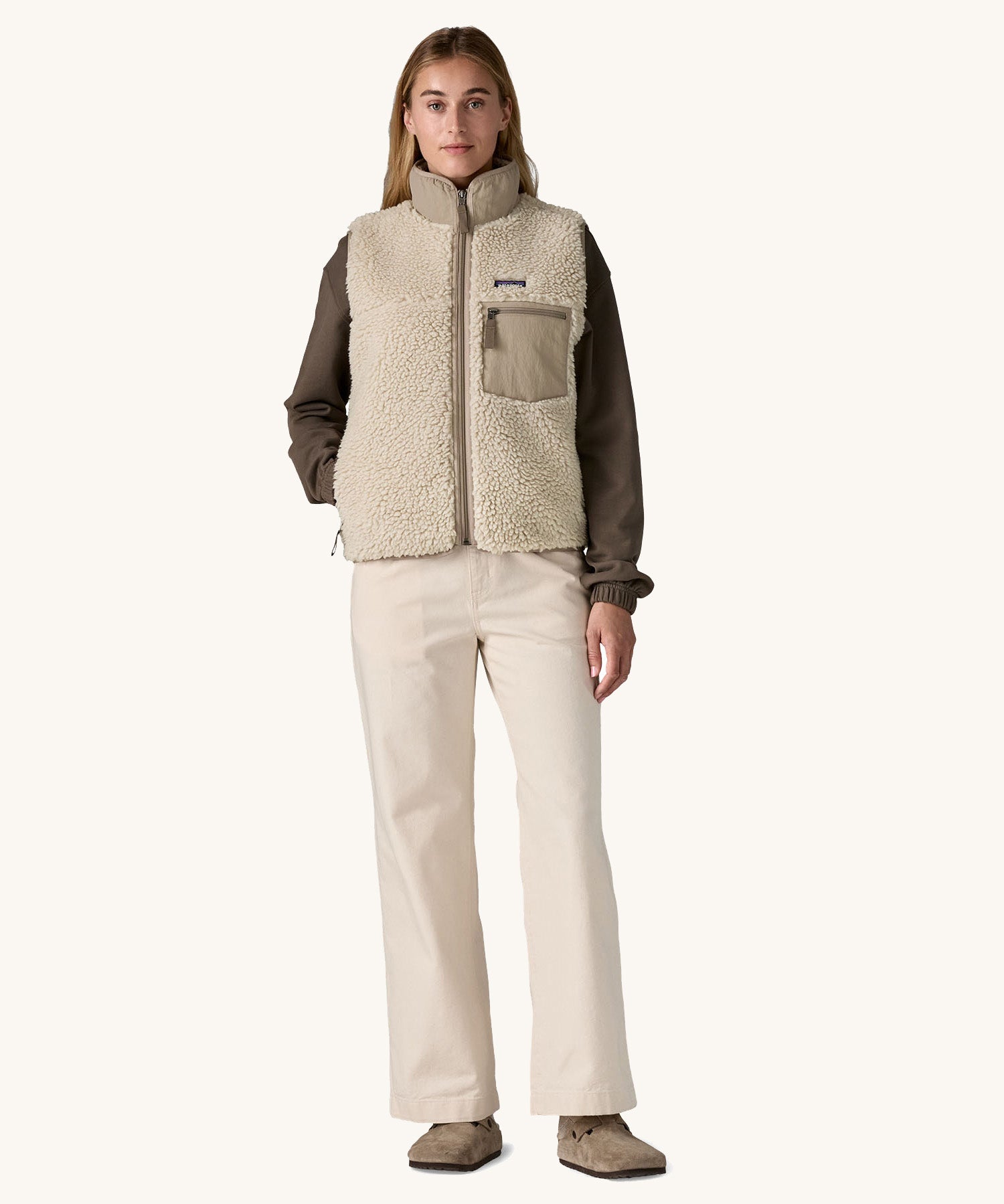 A female standing in front of a cream background and wearing a brown jumper and light cream trousers with the Patagonia thick cream fleece gilet with the zip fully fastened and has a hand in the side pocket