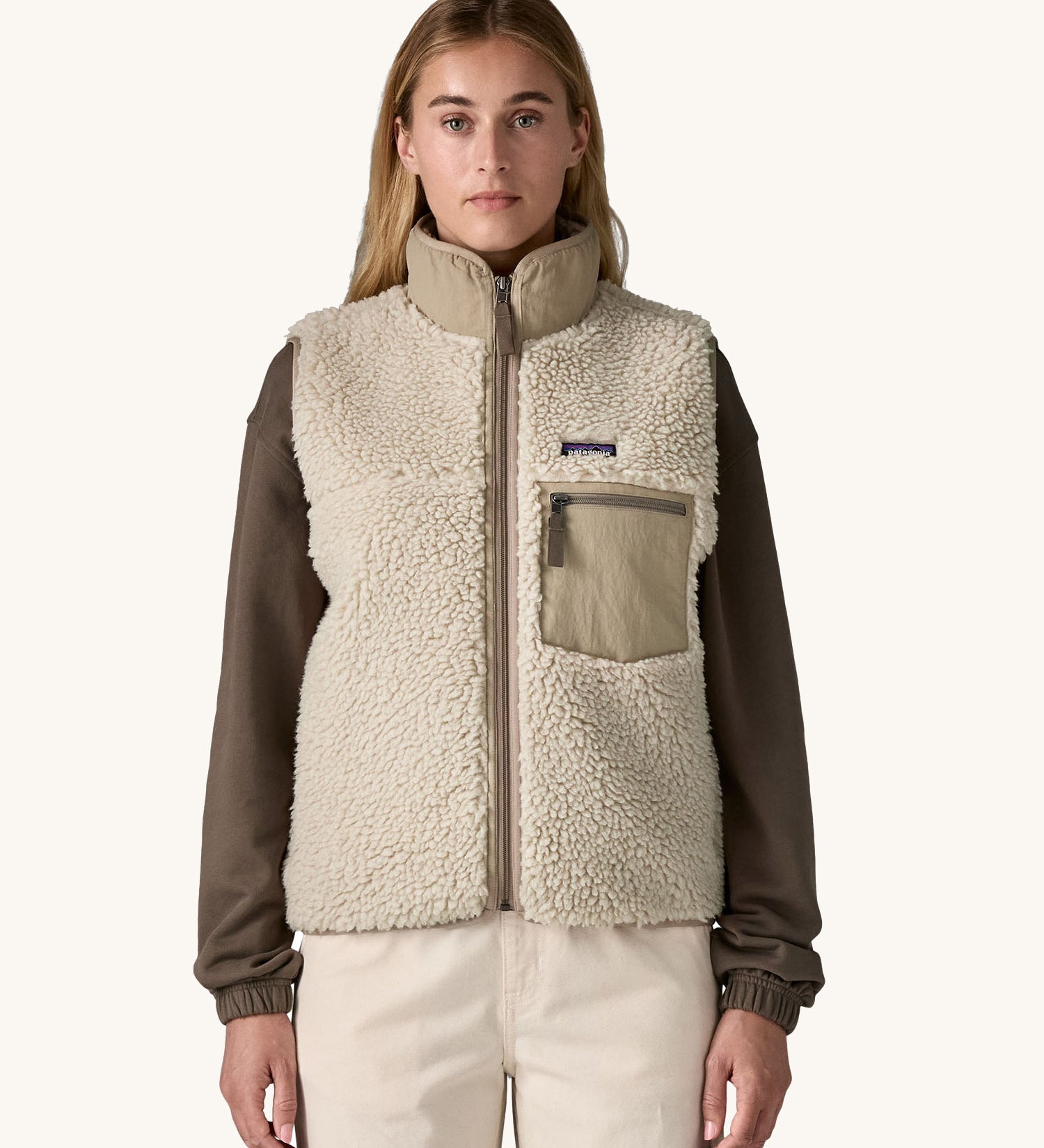 A female standing in front of a cream background and wearing the Patagonia thick cream fleece gilet with the zip fully fastened.