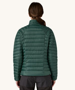 The back of the female standing in front of a cream background and wearing black trousers with the Patagonia puffer style jacket in dark green