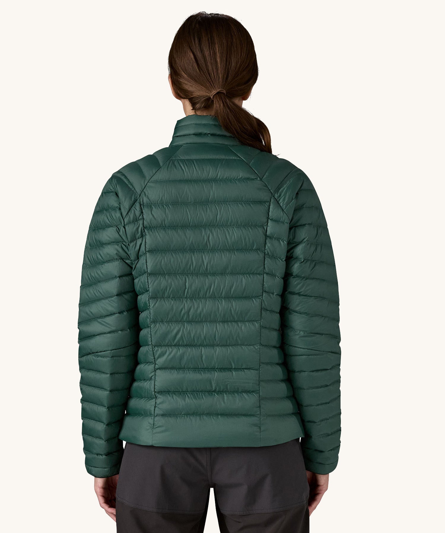 The back of the female standing in front of a cream background and wearing black trousers with the Patagonia puffer style jacket in dark green