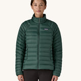 A female standing in front of a cream background and wearing black trousers with the Patagonia puffer style jacket in dark green with the zip fully fastened