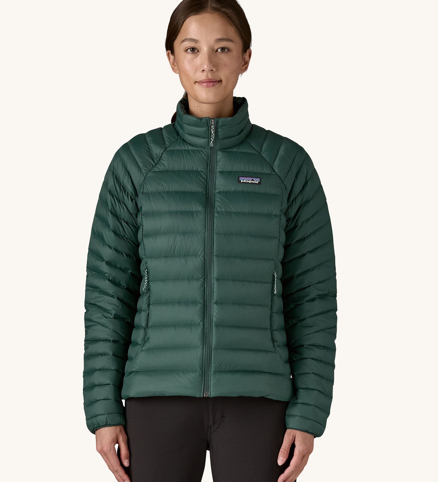 A female standing in front of a cream background and wearing black trousers with the Patagonia puffer style jacket in dark green with the zip fully fastened