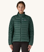 A female standing in front of a cream background and wearing black trousers with the Patagonia puffer style jacket in dark green with the zip fully fastened
