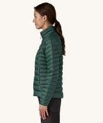 The side of the female standing in front of a cream background and wearing black trousers with the Patagonia puffer jacket in dark green with the zip fully fastened