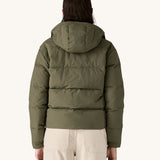 The back of the A female standing in front of a cream background and wearing the Patagonia jacket in dark green with the hood down 