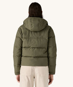 The back of the A female standing in front of a cream background and wearing the Patagonia jacket in dark green with the hood down 