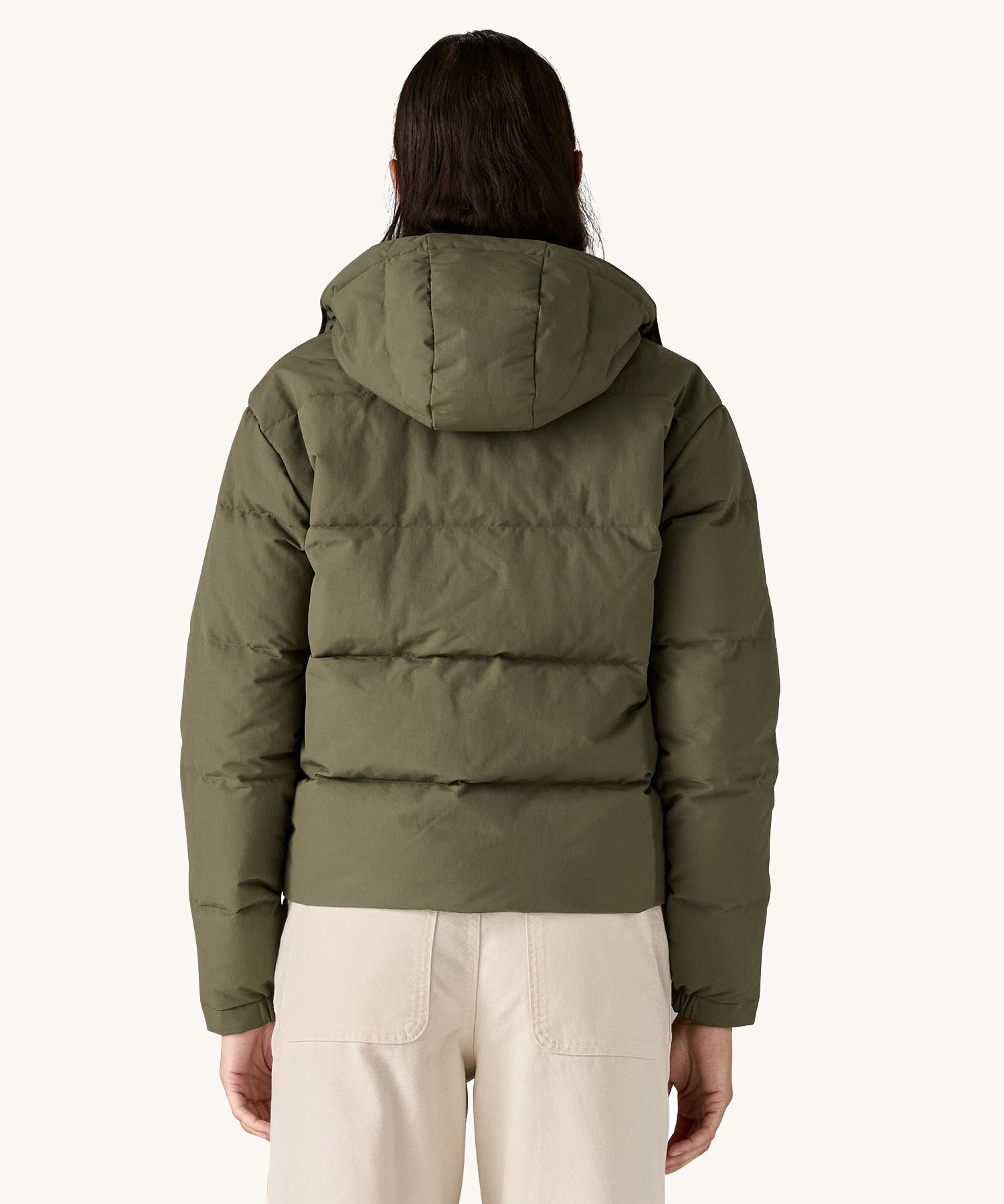 The back of the A female standing in front of a cream background and wearing the Patagonia jacket in dark green with the hood down 