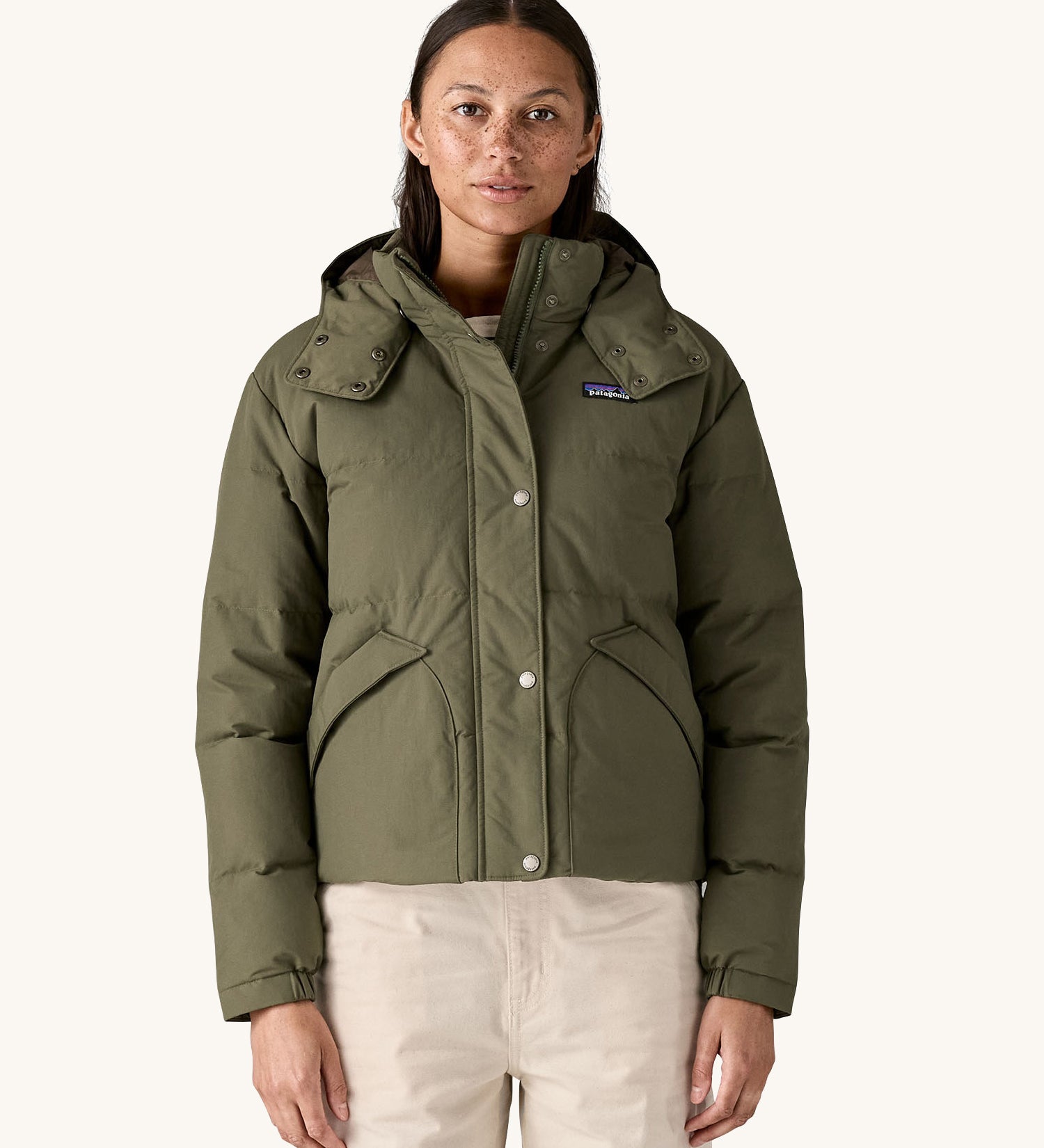 A female standing in front of a cream background and wearing the Patagonia jacket in dark green with the hood down and zip fully fastened.
