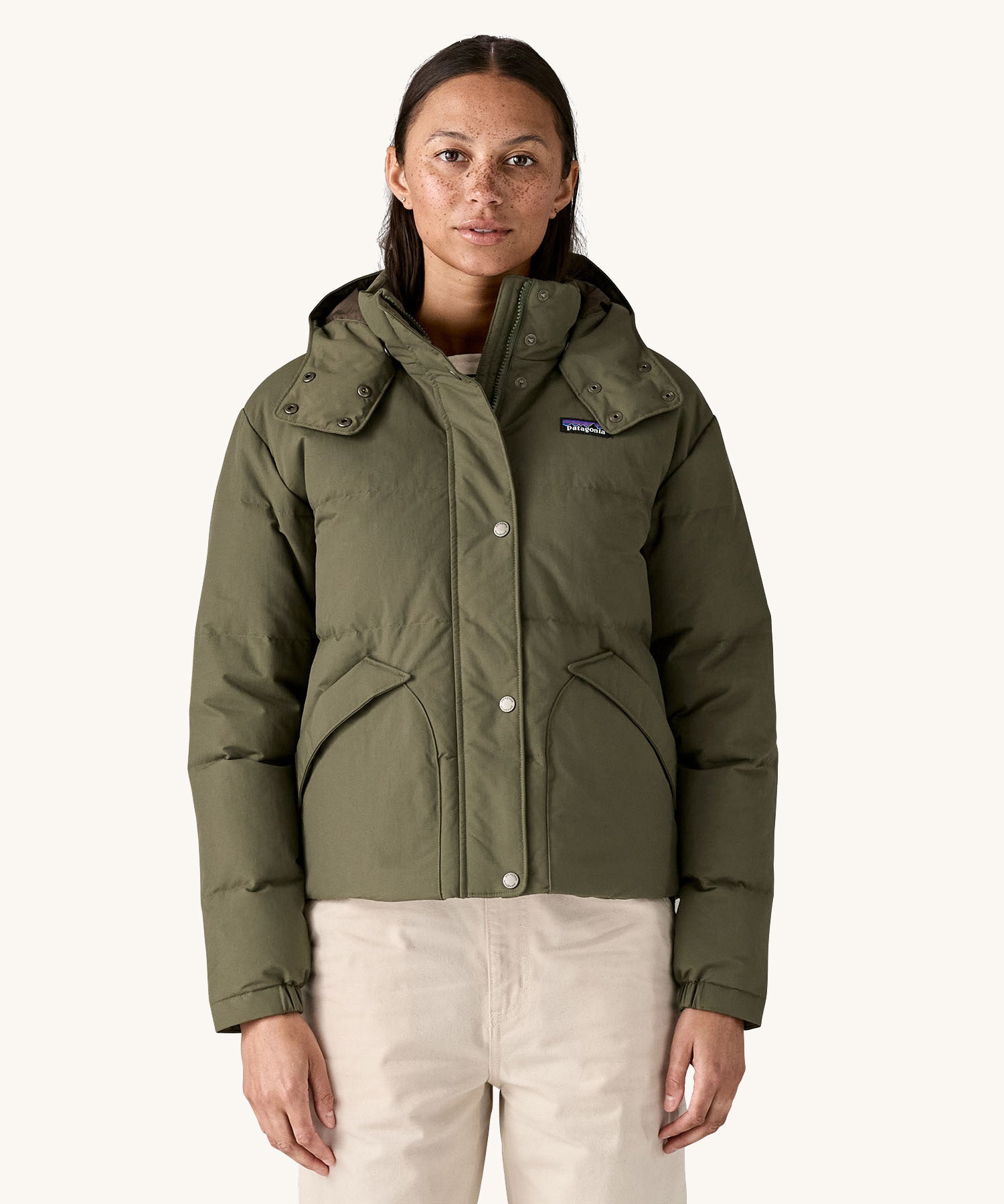 A female standing in front of a cream background and wearing the Patagonia jacket in dark green with the hood down and zip fully fastened.
