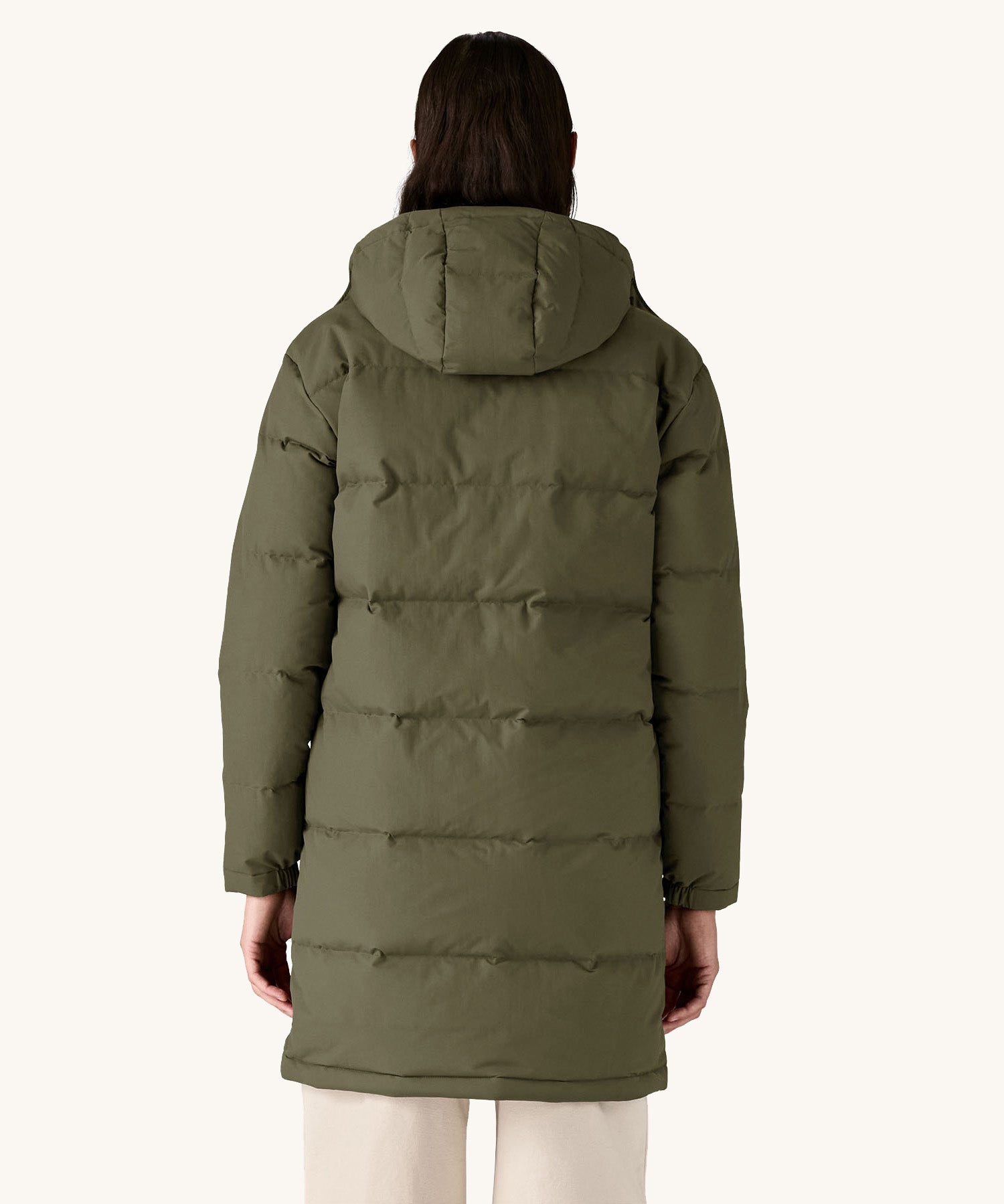 The back of the female model standing in front of a cream background and wearing the Patagonia dark green hooded parka jacket 