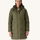 A female standing in front of a cream background and wearing the Patagonia women's dark green hooded parka jacket with side pockets and a popper with zip fastener from the neckline down.