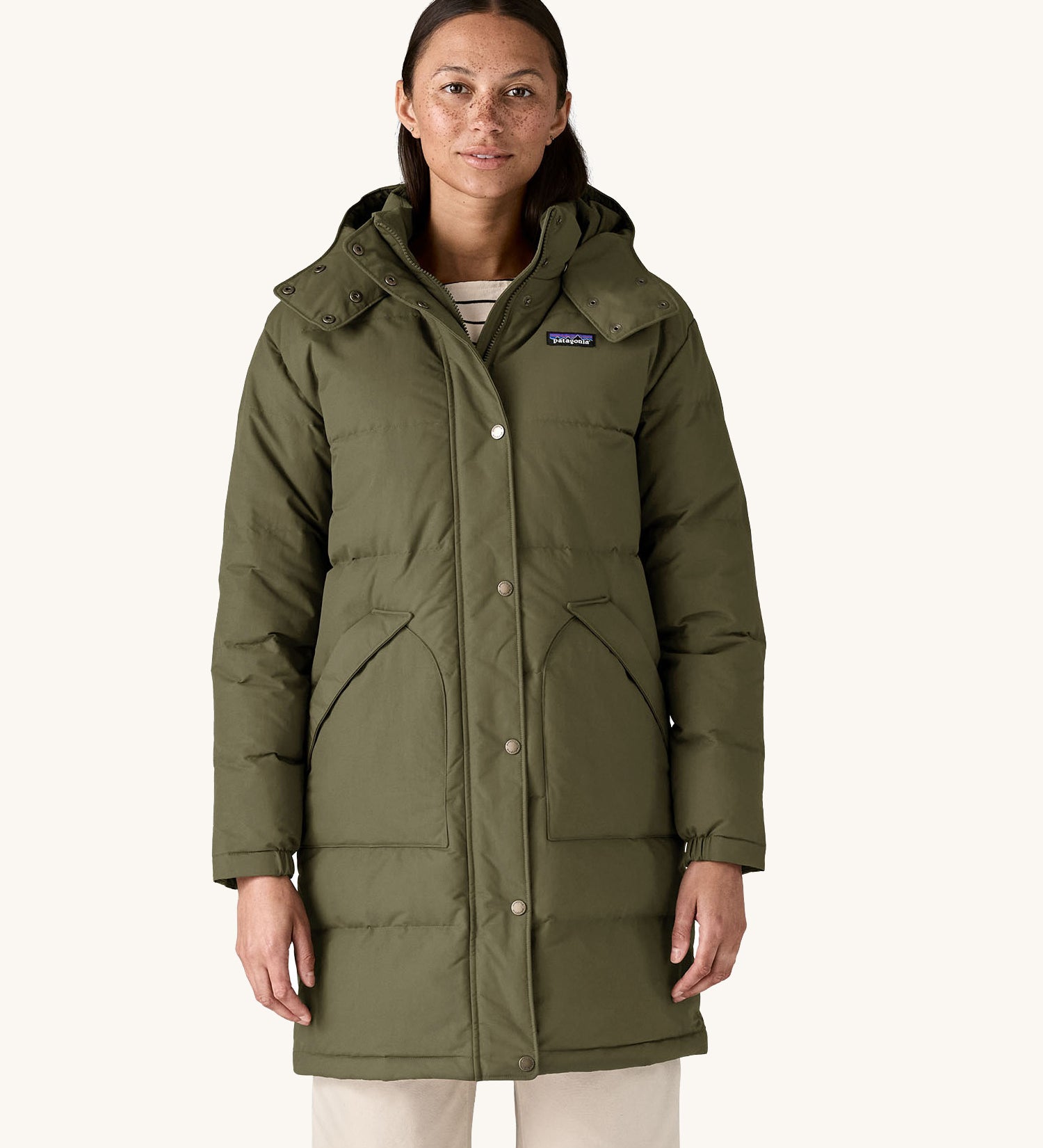 A female standing in front of a cream background and wearing the Patagonia women's dark green hooded parka jacket with side pockets and a popper with zip fastener from the neckline down.