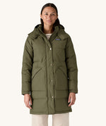 A female standing in front of a cream background and wearing the Patagonia women's dark green hooded parka jacket with side pockets and a popper with zip fastener from the neckline down.