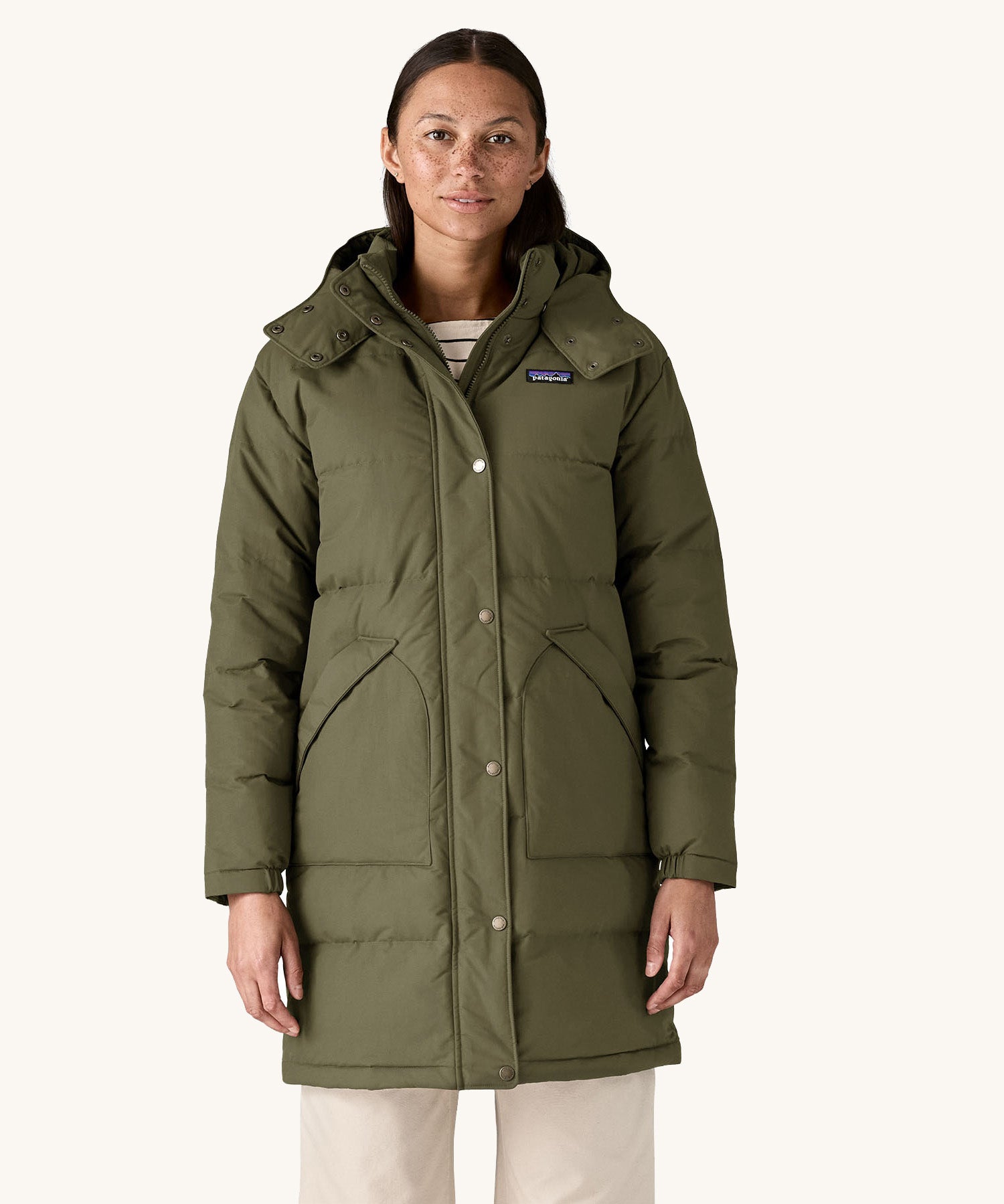 A female standing in front of a cream background and wearing the Patagonia women's dark green hooded parka jacket with side pockets and a popper with zip fastener from the neckline down.