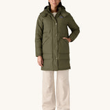 A female standing in front of a cream background and wearing the Patagonia women's dark green hooded parka jacket with a hand in one of the side pockets 