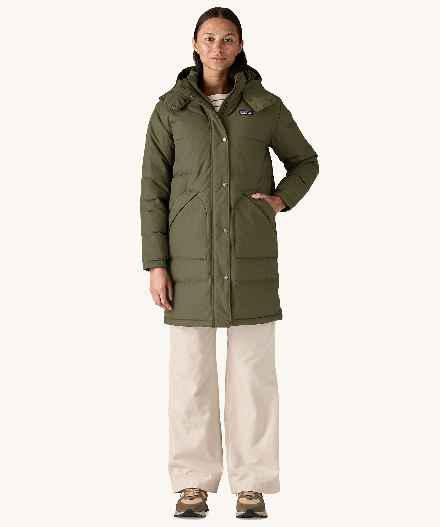 A female standing in front of a cream background and wearing the Patagonia women's dark green hooded parka jacket with a hand in one of the side pockets 