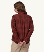 The back of the Female standing in front of a cream background and wearing cream corduroy trousers and the Patagonia brushed cotton shirt in red with a dark red check design.