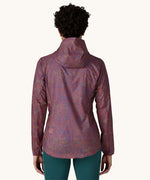 The back of a female standing in front of a cream background wearing dark green leggings and the Patagonia women's lightweight hooded rain jacket in dark red with a repeating purple pattern and the hood down