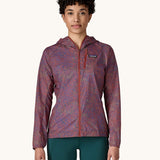 A female standing in front of a cream background wearing dark green leggings and the Patagonia women's lightweight hooded rain jacket in dark red with a repeating purple pattern with the front zip fully fastened