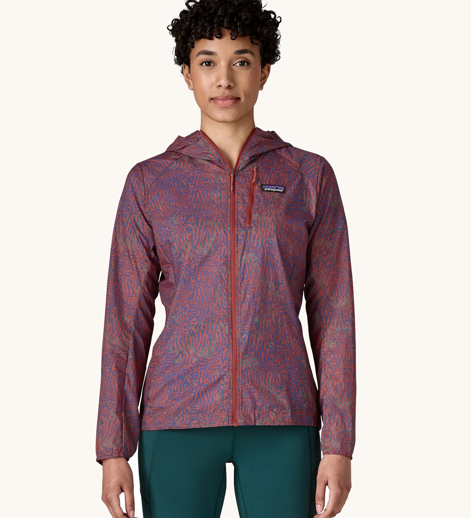 A female standing in front of a cream background wearing dark green leggings and the Patagonia women's lightweight hooded rain jacket in dark red with a repeating purple pattern with the front zip fully fastened