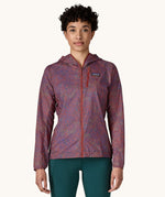 A female standing in front of a cream background wearing dark green leggings and the Patagonia women's lightweight hooded rain jacket in dark red with a repeating purple pattern with the front zip fully fastened