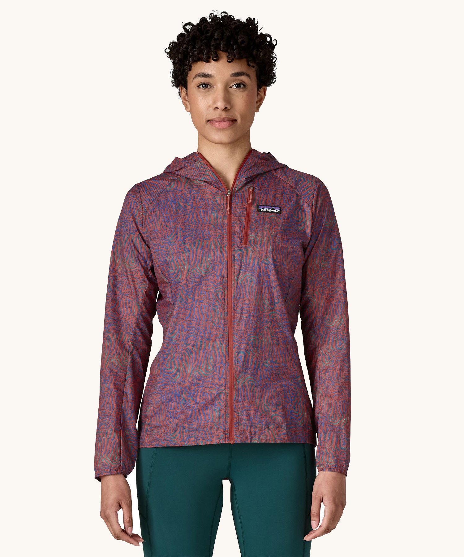 A female standing in front of a cream background wearing dark green leggings and the Patagonia women's lightweight hooded rain jacket in dark red with a repeating purple pattern with the front zip fully fastened