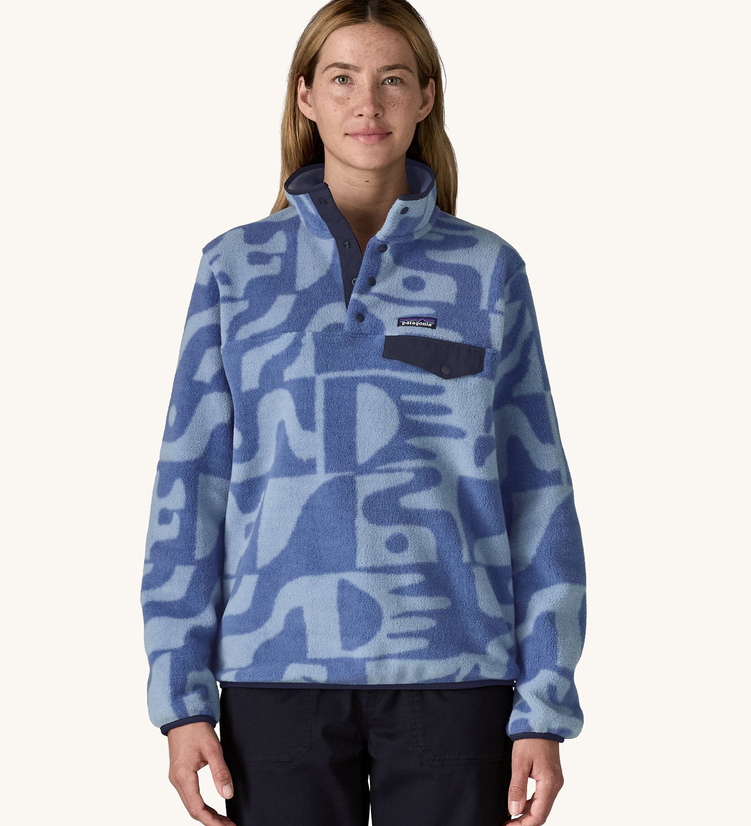 A female standing in front of a cream background. Wearing black trousers and the Patagonia women's lightweight fleece with light and dark blue retro patterns. 