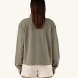 The back of a female standing in front of a cream background, wearing white shorts and the Patagonia women's lightweight fleece in dark green. 
