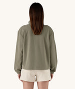 The back of a female standing in front of a cream background, wearing white shorts and the Patagonia women's lightweight fleece in dark green. 