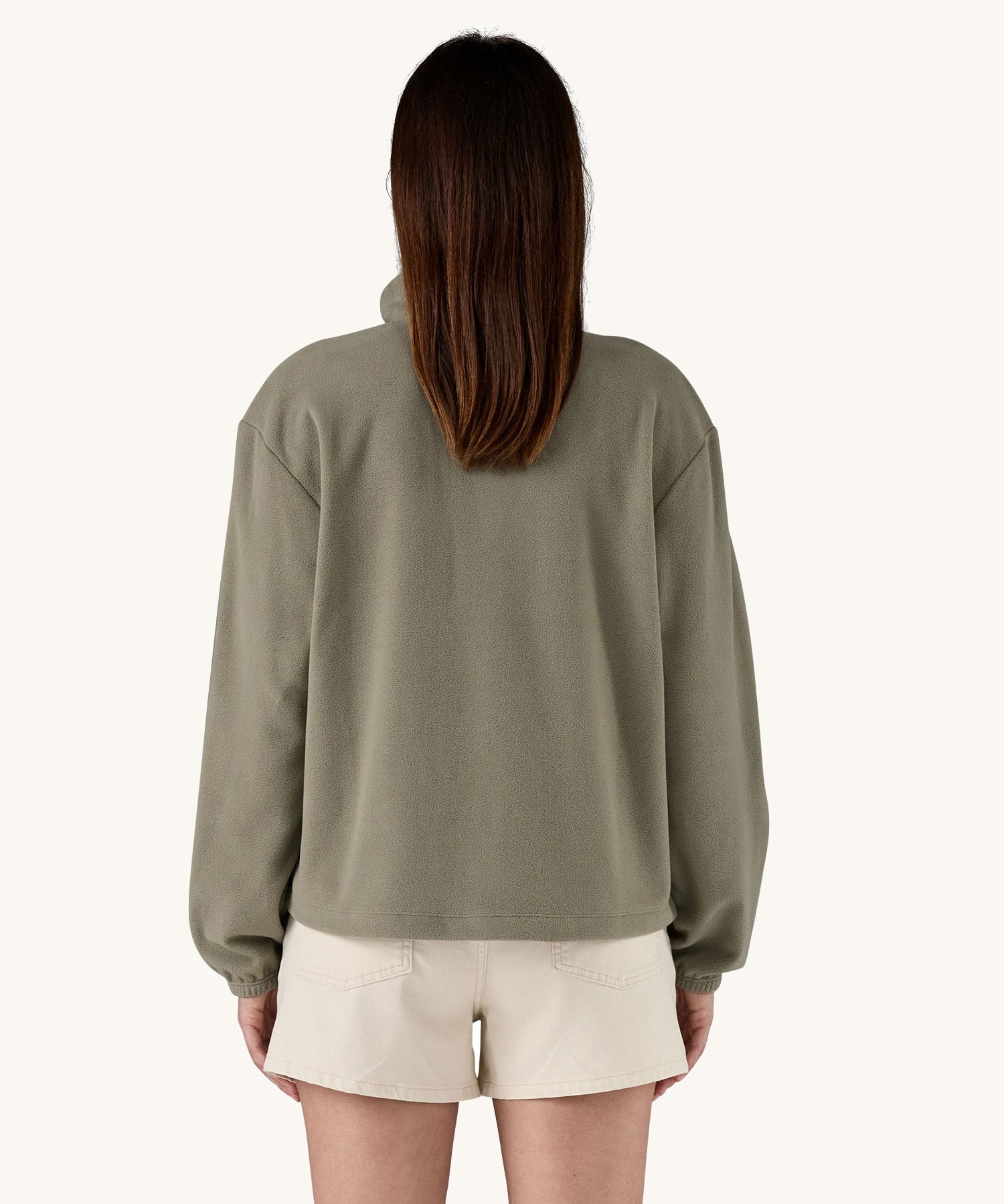 The back of a female standing in front of a cream background, wearing white shorts and the Patagonia women's lightweight fleece in dark green. 
