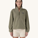 A female standing in front of a cream background, wearing white shorts and the Patagonia women's lightweight fleece in dark green. with the half length front zip fastened