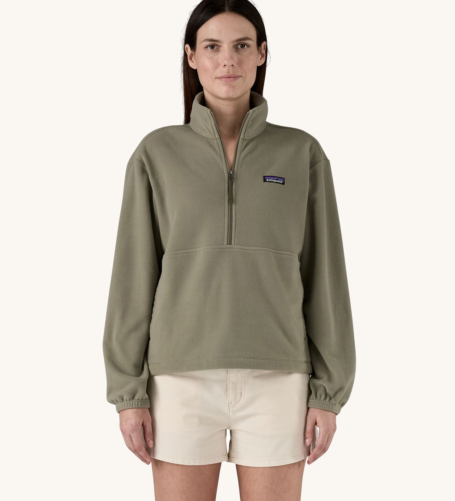 A female standing in front of a cream background, wearing white shorts and the Patagonia women's lightweight fleece in dark green. with the half length front zip fastened