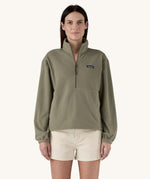 A female standing in front of a cream background, wearing white shorts and the Patagonia women's lightweight fleece in dark green. with the half length front zip fastened