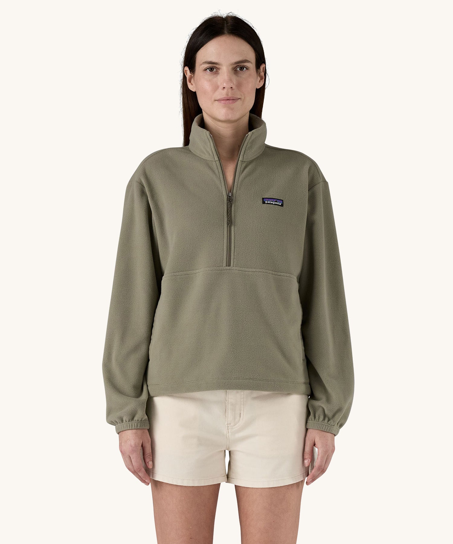 A female standing in front of a cream background, wearing white shorts and the Patagonia women's lightweight fleece in dark green. with the half length front zip fastened