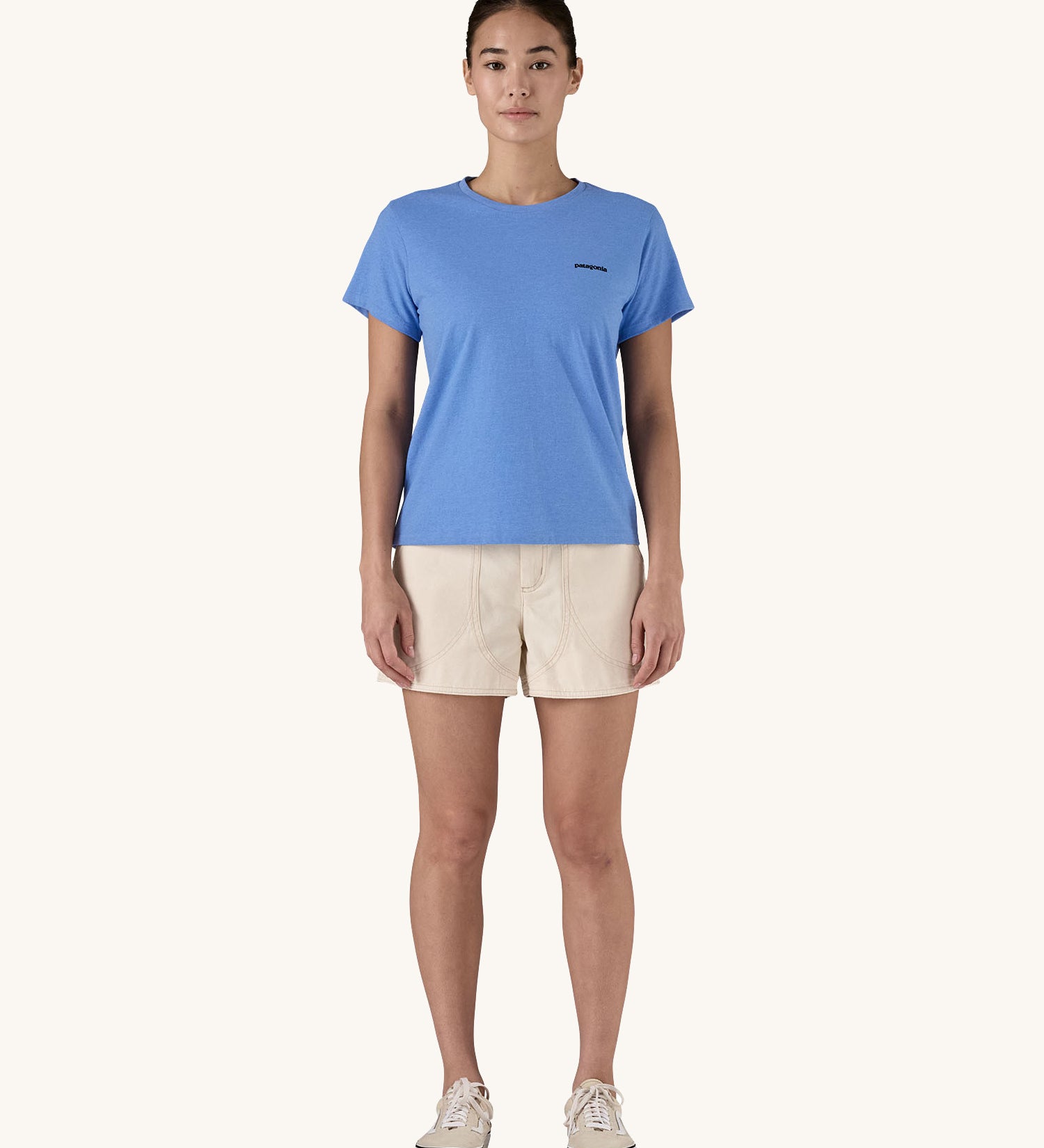 Female standing in front of a cream background wearing cream shorts, trainers and the Patagonia organic cotton short sleeve t-shirt in light blue 