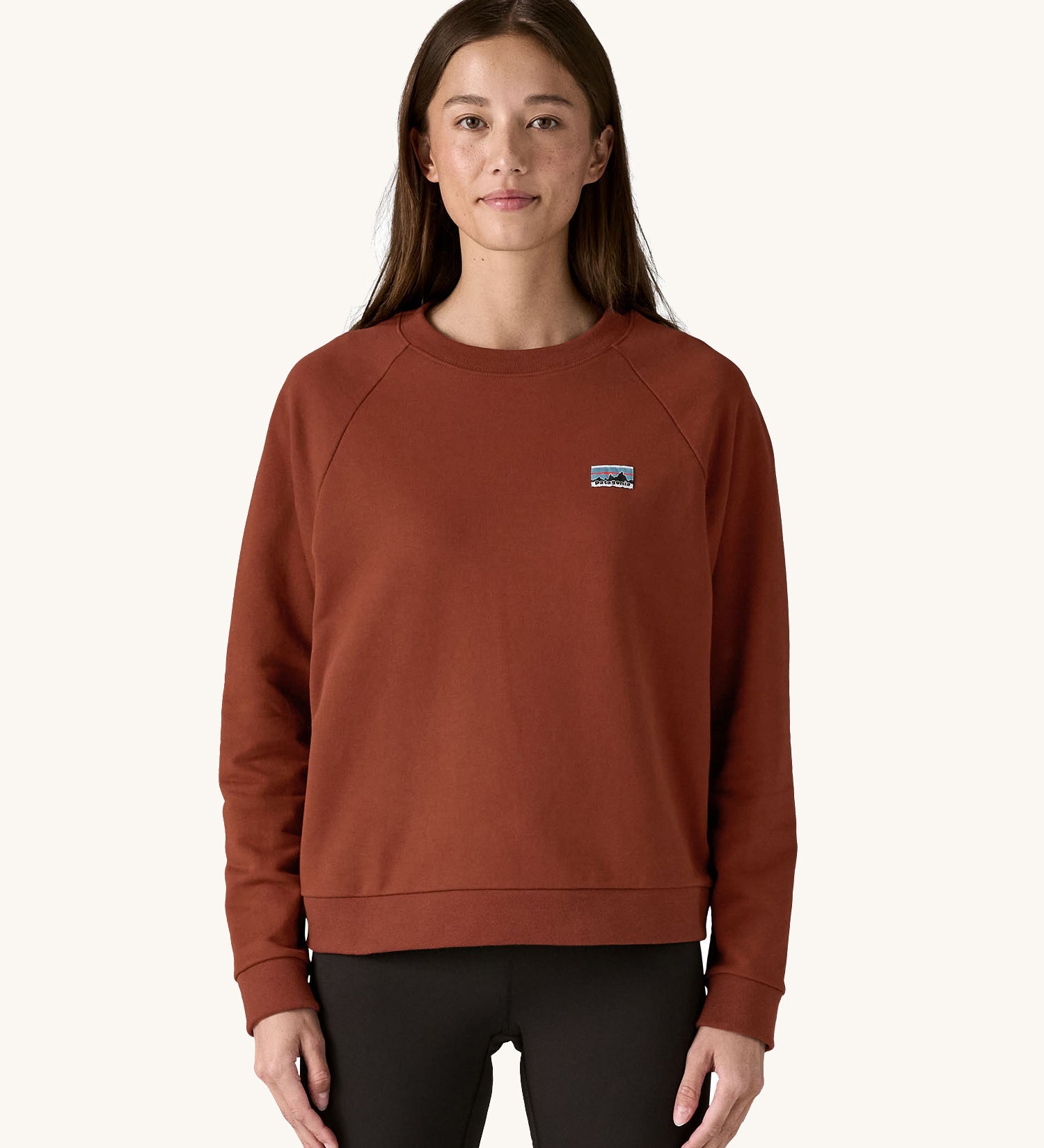 A female standing in front of a cream background and wearing black leggings with the Patagonia women's organic cotton sweater in dark red with a small Patagonia patch on the chest