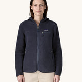 A female standing in front of a cream background and wearing the dark blue Patagonia thick hooded fleece with the zip fully fastened 