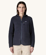 A female standing in front of a cream background and wearing the dark blue Patagonia thick hooded fleece with the zip fully fastened 