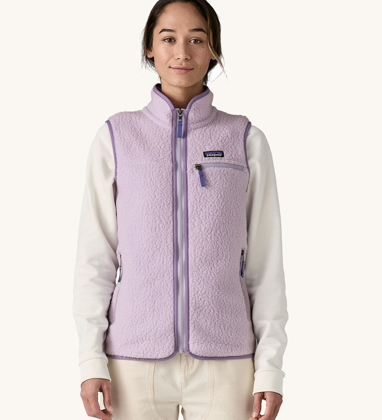 A female standing in front of a cream background and wearing the light purple Patagonia women's thick fleece gilet with the zip fully closed