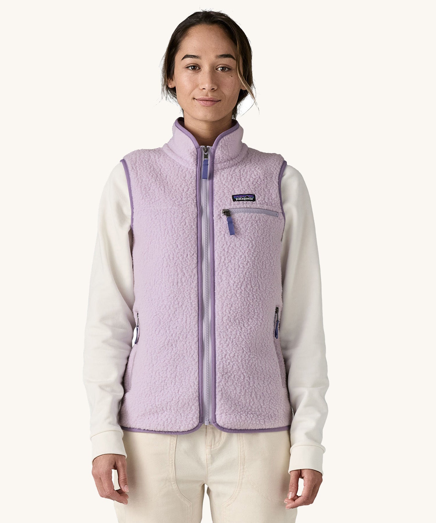 A female standing in front of a cream background and wearing the light purple Patagonia women's thick fleece gilet with the zip fully closed