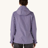 The back of the female standing in front of a cream background and wearing dark grey walking trousers with the Patagonia rain coat in pastel purple colour with the hood down