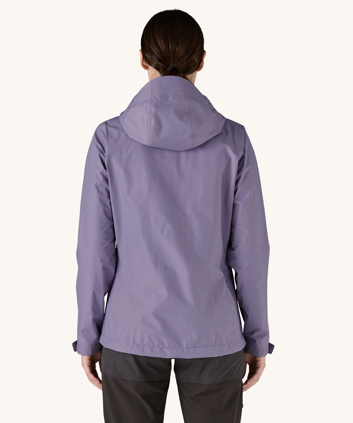 The back of the female standing in front of a cream background and wearing dark grey walking trousers with the Patagonia rain coat in pastel purple colour with the hood down