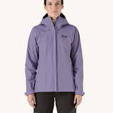 A female standing in front of a cream background and wearing dark grey walking trousers with the Patagonia hooded rain coat in pastel purple colour with a full length front zip and side pockets.