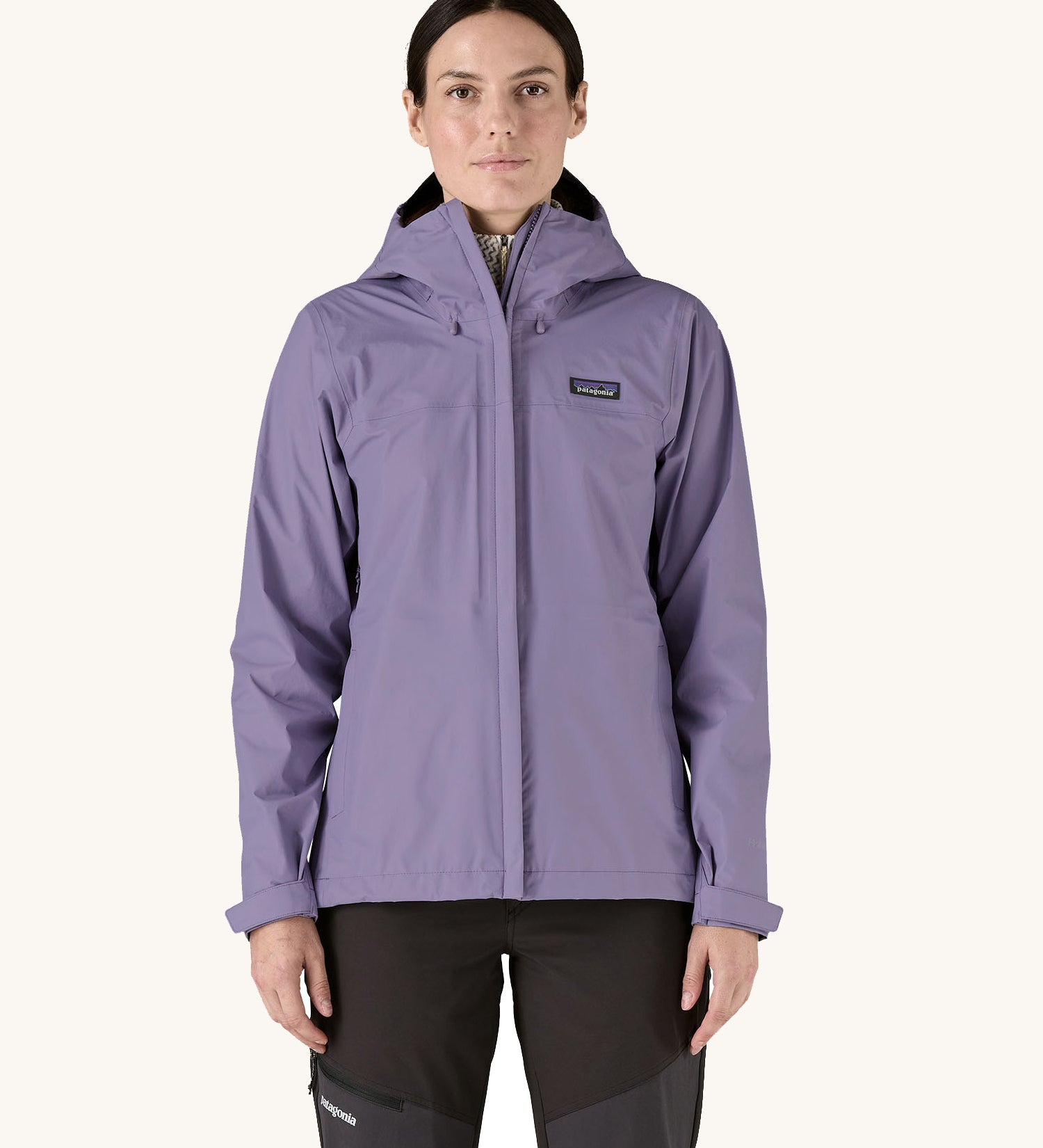 A female standing in front of a cream background and wearing dark grey walking trousers with the Patagonia hooded rain coat in pastel purple colour with a full length front zip and side pockets.