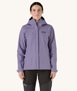 A female standing in front of a cream background and wearing dark grey walking trousers with the Patagonia hooded rain coat in pastel purple colour with a full length front zip and side pockets.