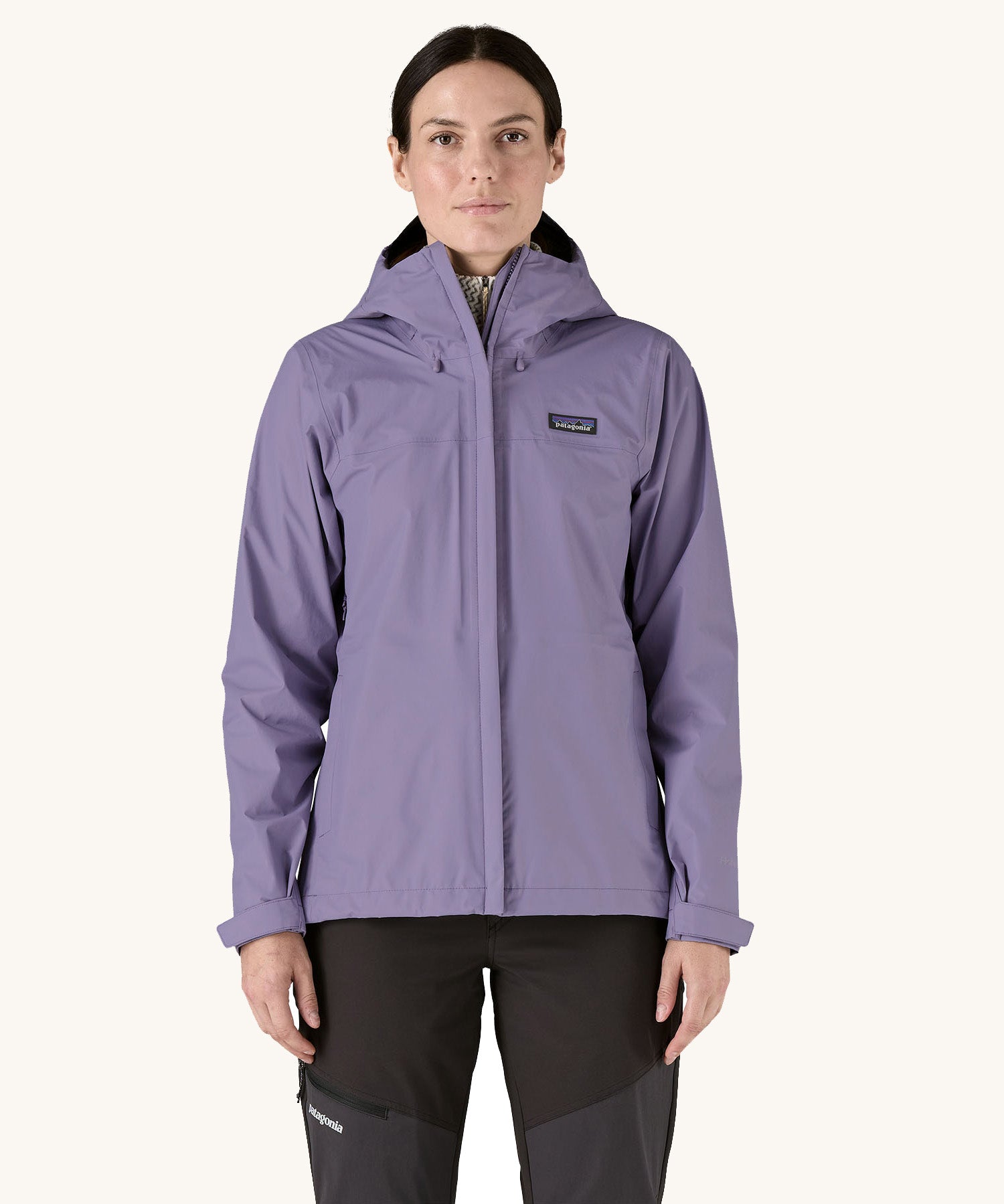 A female standing in front of a cream background and wearing dark grey walking trousers with the Patagonia hooded rain coat in pastel purple colour with a full length front zip and side pockets.