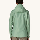 The back of the female standing in front of a cream background and wearing brown walking trousers with the Patagonia rain coat in pastel green colour with the hood down