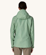 The back of the female standing in front of a cream background and wearing brown walking trousers with the Patagonia rain coat in pastel green colour with the hood down