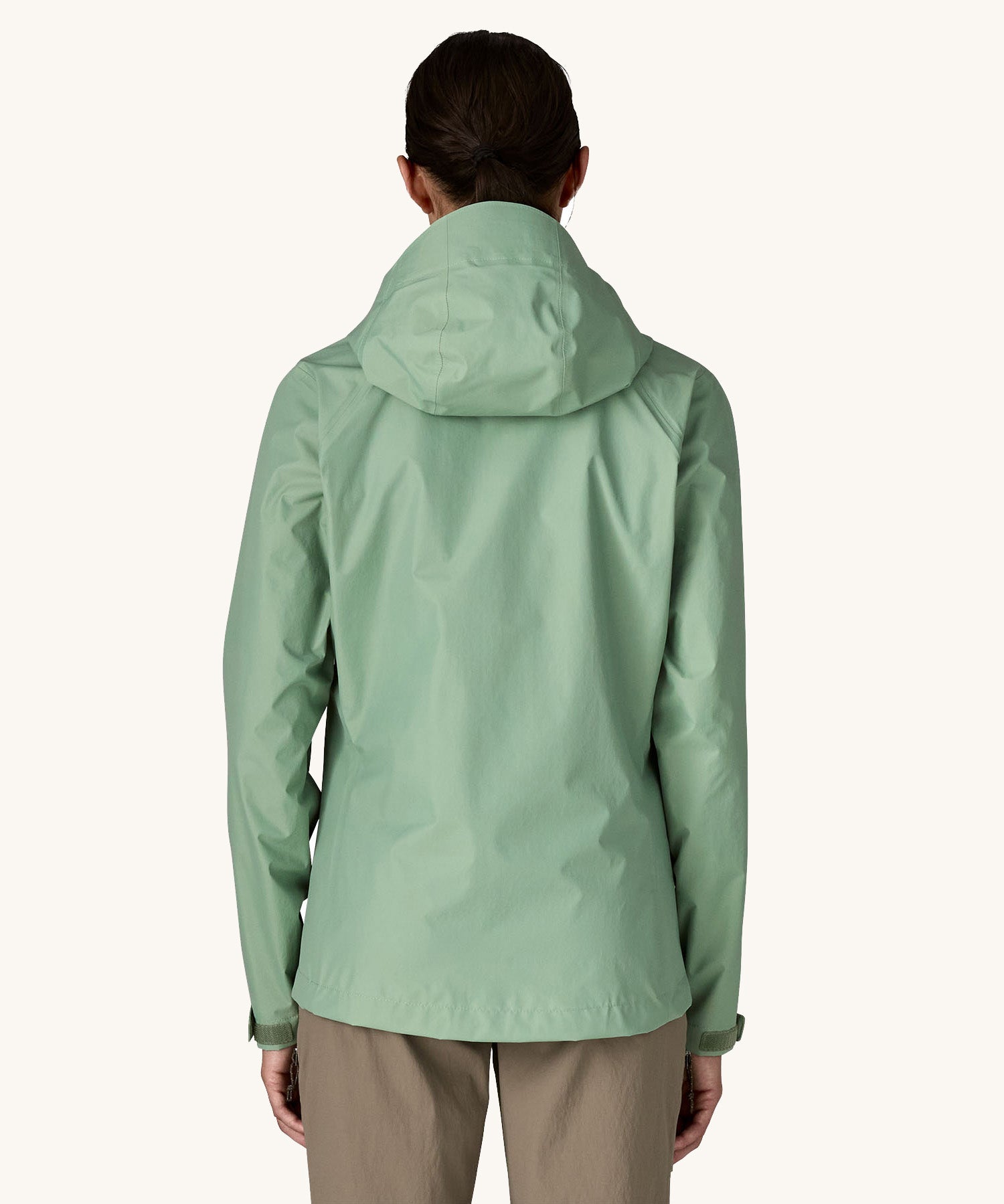 The back of the female standing in front of a cream background and wearing brown walking trousers with the Patagonia rain coat in pastel green colour with the hood down