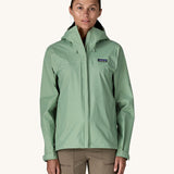 A female standing in front of a cream background and wearing brown walking trousers with the Patagonia hooded rain coat in pastel green colour with a full length front zip and side pockets.